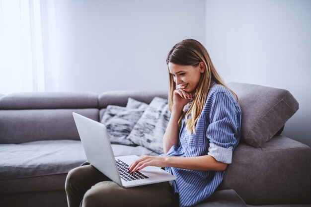 Młoda atrakcyjna uśmiechnięta caucasian caucasian brunetka siedzi na kanapie w żywym pokoju i używa laptop dla freelance pracy.