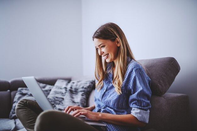 Młoda atrakcyjna uśmiechnięta caucasian caucasian brunetka siedzi na kanapie w żywym pokoju i używa laptop dla freelance pracy.