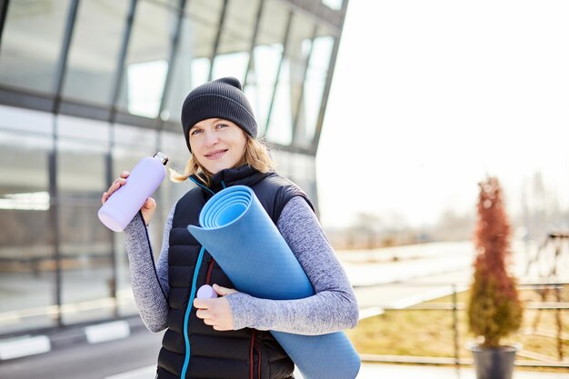 Młoda atrakcyjna uśmiechnięta kobieta uprawiająca sport w porannym wschodzie słońca w parku trzymająca matę do jogi i butelkę wody zdrowy styl życia ubrana w czarny bez rękawów