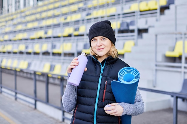 Młoda atrakcyjna uśmiechnięta kobieta uprawiająca sport w porannym wschodzie słońca w parku trzymająca matę do jogi i butelkę wody zdrowy styl życia ubrana w czarny bez rękawów