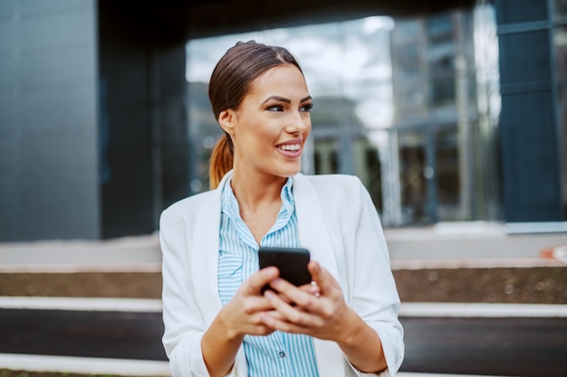 Młoda atrakcyjna uśmiechnięta stylowa bizneswoman stoi przed firmą korporacyjną, trzymając inteligentny telefon i odwracając wzrok.