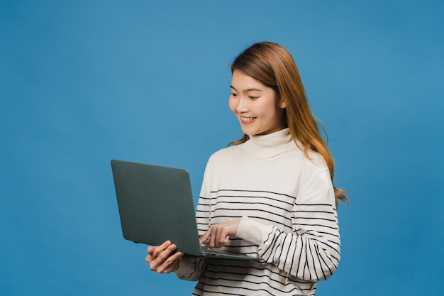 Zdjęcie młoda azjatka korzystająca z laptopa z pozytywną ekspresją, uśmiecha się szeroko, ubrana w zwykłe ubranie, czująca szczęście i stojąca na białym tle na niebieskiej ścianie