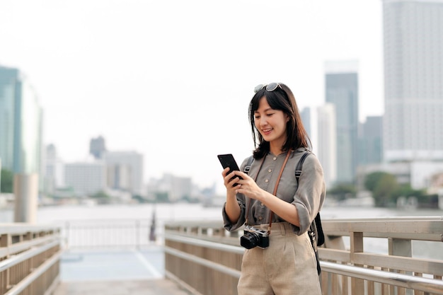 Młoda Azjatka podróżująca z plecakiem za pomocą telefonu komórkowego na molo ekspresowej łodzi na rzece Chao Phraya w Bangkoku