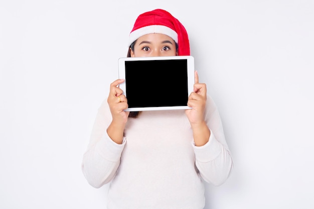 Młoda Azjatka w kapeluszu Boże Narodzenie zakrywająca twarz na tablecie z pustym ekranem, izolowana na białym tle Indonezyjczycy świętują koncepcję Bożego Narodzenia