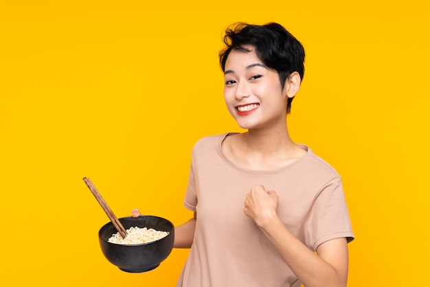 Młoda Azjatycka dziewczyna nad odosobnioną kolor żółty ścianą z niespodzianka wyrazem twarzy podczas gdy trzymający puchar kluski z chopsticks