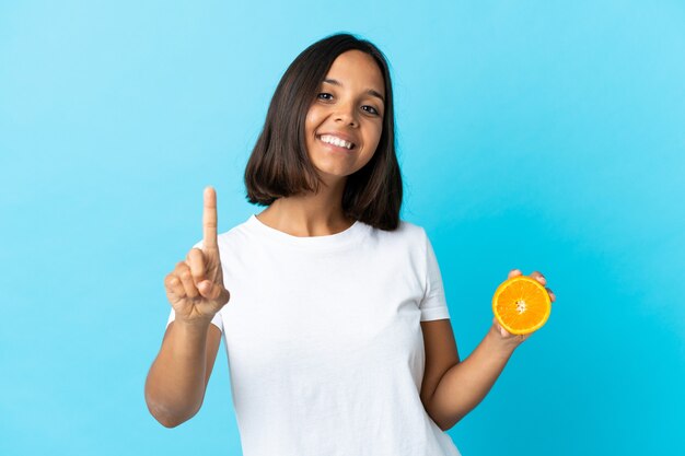 Młoda azjatycka dziewczyna trzyma pomarańczę na białym tle na niebieskim tle pokazując i podnosząc palec