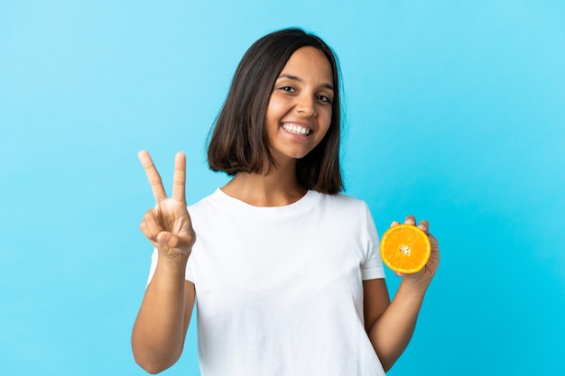Młoda azjatycka dziewczyna trzyma pomarańczowy na białym tle na niebieskiej ścianie, uśmiechając się i pokazując znak zwycięstwa