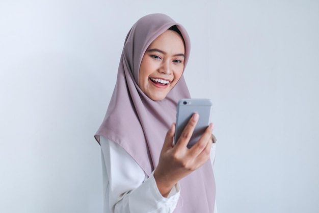 Młoda azjatycka islamska kobieta nosząca chustę jest szczęśliwa i podekscytowana świętując to, co widzi na smartfonie Indonezyjska kobieta na szarym tle