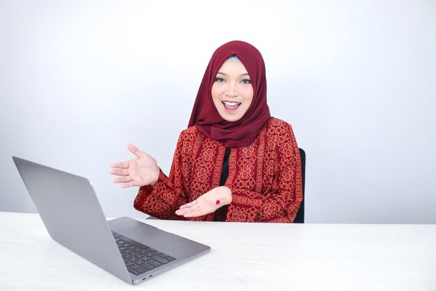 Młoda azjatycka islamska kobieta uśmiecha się wskazując ręką podczas pracy na laptopie na białym tle