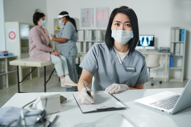 Młoda azjatycka klinicysta w ochronnej odzieży roboczej siedzi przy biurku i wypełnia dokument, podczas gdy jej kolega szczepi pacjenta w tle