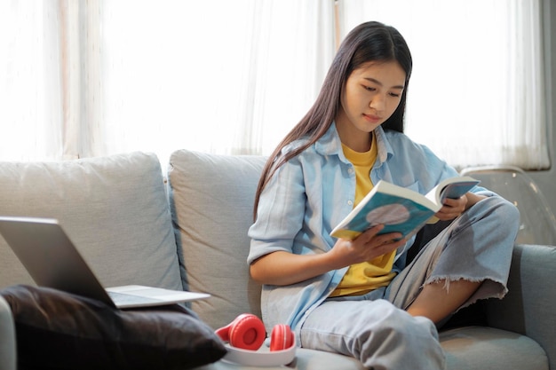 Młoda azjatycka kobieta czytająca książkę przed laptopem w domu