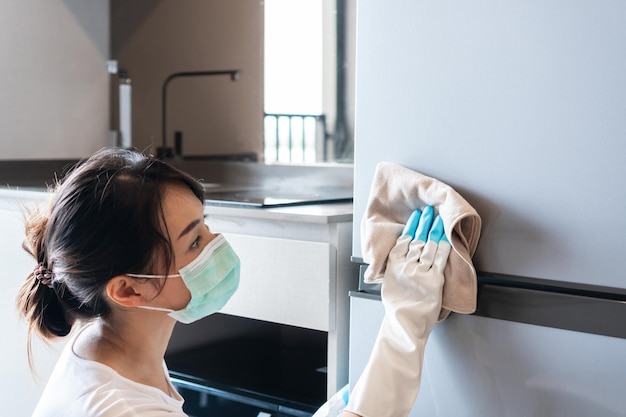 Młoda Azjatycka kobieta nosi maskę medyczną i ręce w gumowej białej rękawiczce do czyszczenia lodówki z serwetką w kuchni w domu Zbliżenie