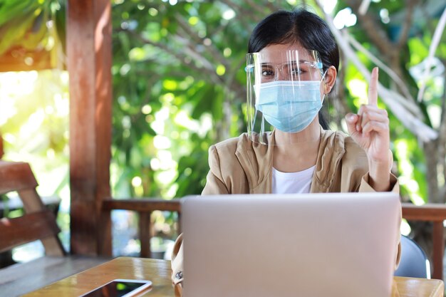 Młoda azjatycka kobieta ręce w casualowej sukience z maską ochronną dla opieki zdrowotnej, siedząca w kawiarni i używająca smartfona i pracująca na komputerze przenośnym. Nowa koncepcja normalnego i społecznego dystansu