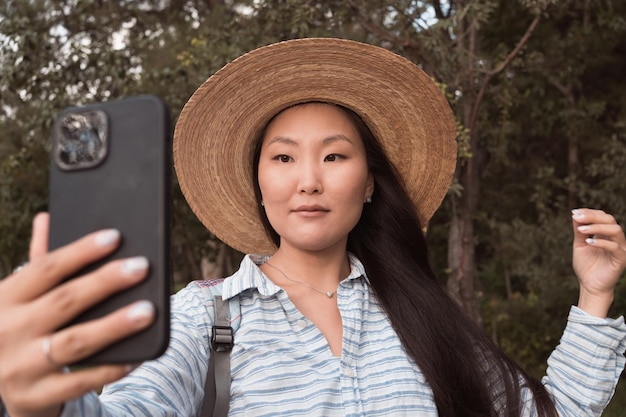 Młoda azjatycka kobieta w kapeluszu robi zdjęcie selfie przez telefon w parku miejskim latem