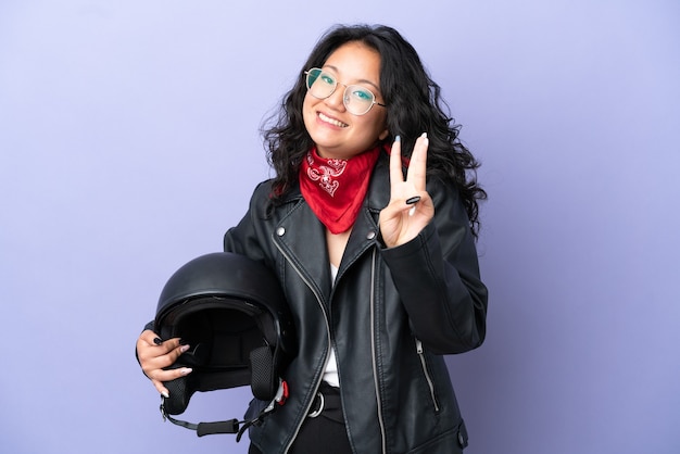 Młoda azjatycka kobieta z kaskiem motocyklowym na fioletowym tle szczęśliwa i licząca trzy palcami