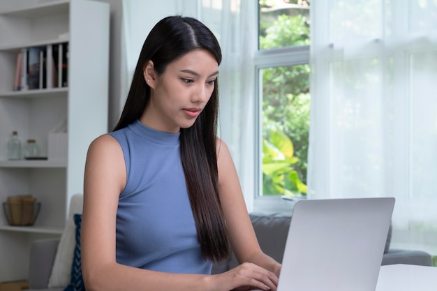Młoda Azjatycka kobieta za pomocą laptopa w domu, patrząc na ekran, rozmawiając, czytając lub pisząc e-mail, siedząc na biurku poważna studentka odrabiająca pracę domową, pracująca nad projektem badawczym online