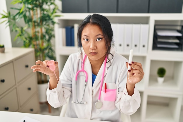 Młoda azjatycka lekarka trzymająca kubek menstruacyjny i tampon, nieświadoma i zdezorientowana. koncepcja wątpliwości.