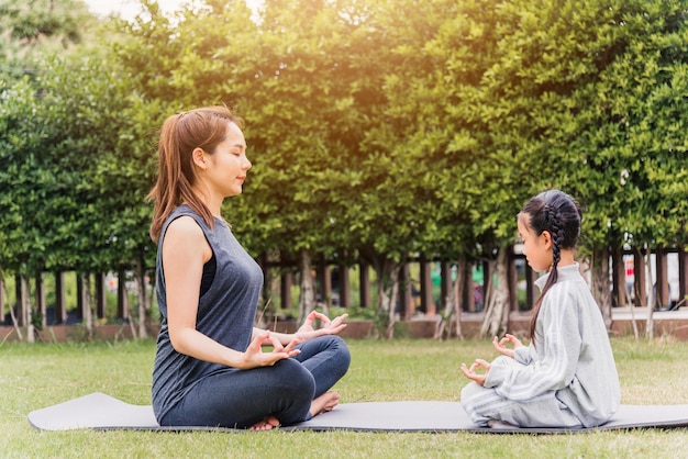 Młoda azjatycka matka ćwiczy ćwiczenia jogi z córką na świeżym powietrzu w medytacyjnej pozie razem na zielonej trawie w przyrodzie w parku ogrodniczym, rodzinnym sporcie i ćwiczeniach dla zdrowego stylu życia