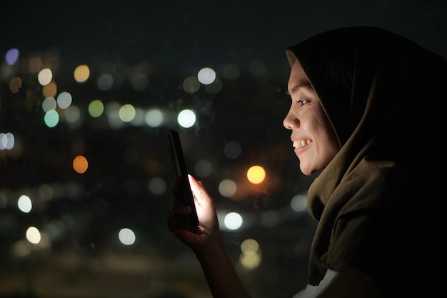 Młoda azjatycka muzułmanka w chustce na głowie leżąca w łóżku i używająca telefonu komórkowego w nocy