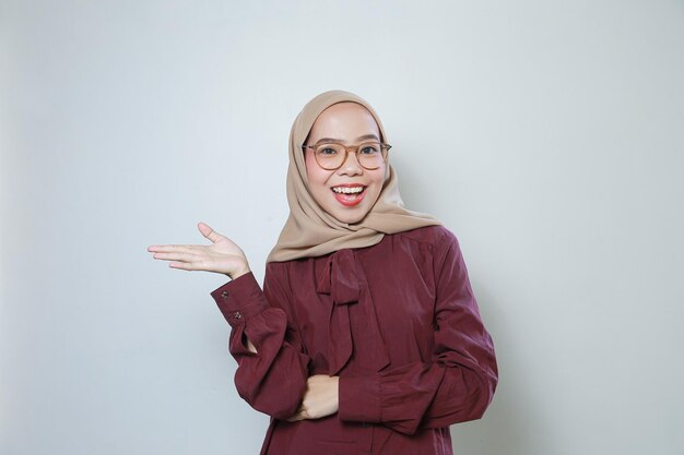 Młoda azjatycka muzułmanka w okularach wskazujących na pustą przestrzeń uśmiecha się, czując się pewnie i szczęśliwie