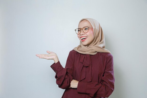 Młoda azjatycka muzułmanka w okularach wskazujących na pustą przestrzeń uśmiecha się, czując się pewnie i szczęśliwie