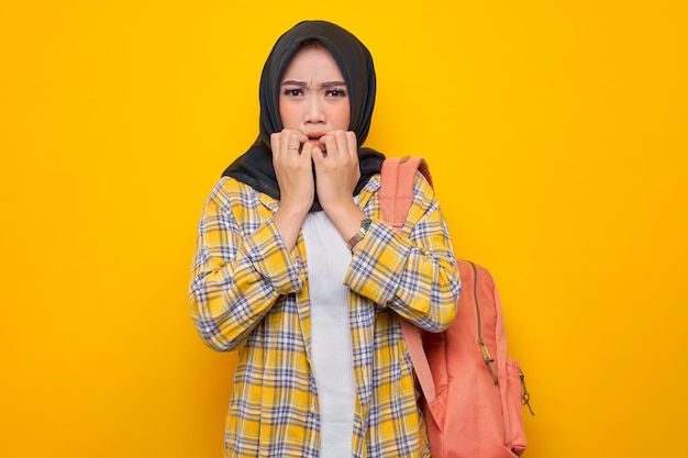 Młoda azjatycka muzułmańska studentka w swobodnych ubraniach i plecaku, wyglądająca na zestresowaną i zdenerwowaną, z ręką na ustach gryzie paznokcie na białym tle na żółtym tle Edukacja szkoła uniwersytet koncepcja kolegium