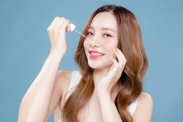 Młoda azjatycka piękna kobieta kręcone długie włosy z koreańskim stylem makijażu na twarzy i idealnie czystą skórą za pomocą kroplomierza na izolowanym niebieskim tle Zabiegi na twarz Kosmetologia Chirurgia plastyczna