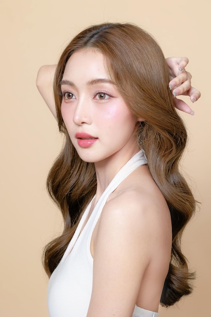 Młoda azjatycka piękna kobieta z kręconymi długimi włosami w koreańskim stylu makijażu dotyka jej twarzy i doskonałej skóry na izolowanym beżowym tle Leczenie twarzy Kosmetologia chirurgia plastyczna