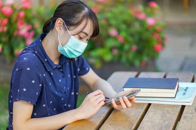 Zdjęcie młoda azjatycka studencka kobieta jest ubranym medyczną maskę i trzyma smartphone, patrzeje ekran, używa app lub przesyłanie wiadomości, podczas gdy siedzący przy ogrodową ławką z książką. nowa normalna koncepcja po covid-19