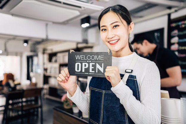 Młoda Azjatycka właścicielka kawiarni barista trzymająca tablicę ogłoszeń "WITAJ", "OPEN" z rozmytym baristą w tle. Biznes z partnerem. Udana praca zespołowa przedsiębiorcy.