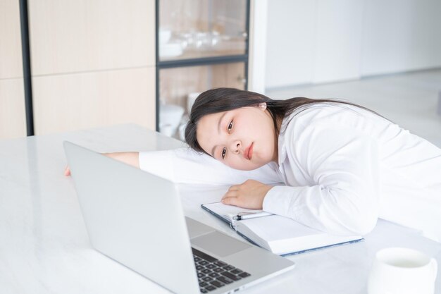 Młoda azjatycka zmęczona biznesowa kobieta z długimi włosami pracuje przy laptopie w jasnym domowym biurze