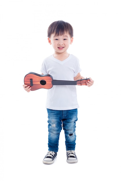 Młoda azjatykcia chłopiec mienia gitary zabawka i uśmiechy nad białym tłem
