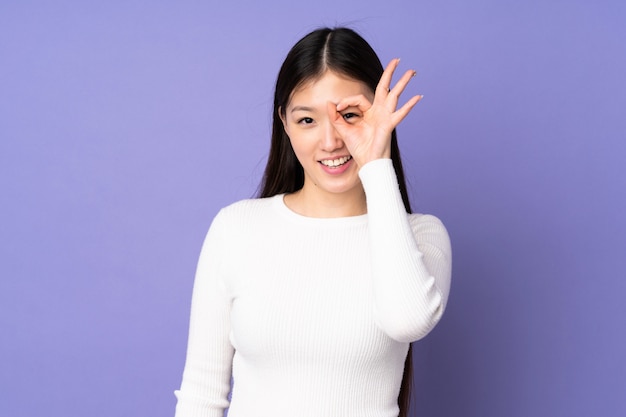 Młoda azjatykcia kobieta odizolowywająca na purpury ścianie pokazuje ok znaka z palcami