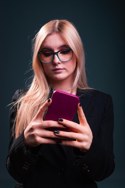 Młoda biznesowa kobieta rasy kaukaskiej korzystająca z telefonu komórkowego, ubrana w czarną marynarkę i okulary w studiu fotograficznym na ciemnym niebieskim tle.