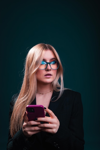 Młoda biznesowa kobieta rasy kaukaskiej korzystająca z telefonu komórkowego, ubrana w czarną marynarkę i okulary w studiu fotograficznym na ciemnym niebieskim tle.