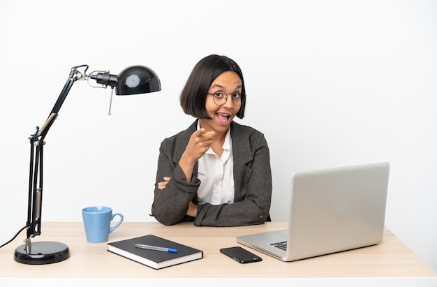Młoda biznesowa kobieta rasy mieszanej pracująca w biurze zaskoczona i wskazująca przód