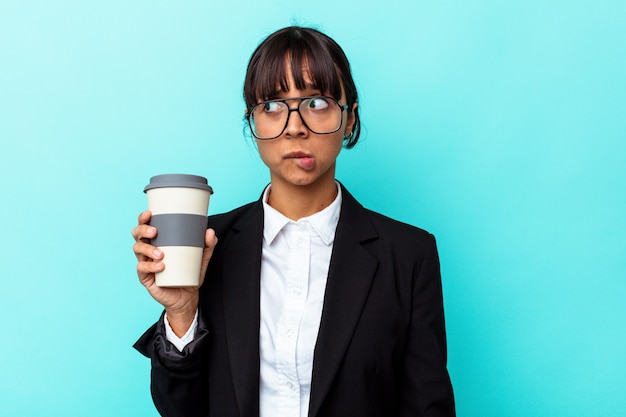 Młoda biznesowa kobieta rasy mieszanej trzyma kawę na białym tle na niebieskim tle zdezorientowana, czuje się wątpliwa i niepewna.