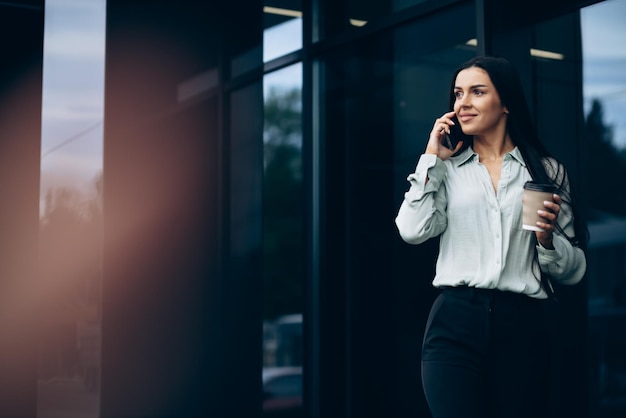 Młoda biznesowa kobieta rozmawia przez telefon i pije kawę przy centrum biznesowym