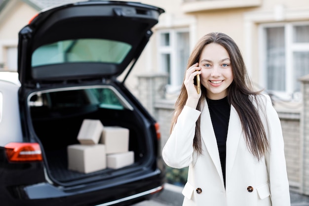 Młoda biznesowa kobieta stojąca przy samochodzie z paczkami wracającymi do domu samochodem Dziewczyna stoi i rozmawia przez telefon komórkowy w pobliżu samochodu Koncepcja kupowania towarów przez Internet i dostarczania ich do domu