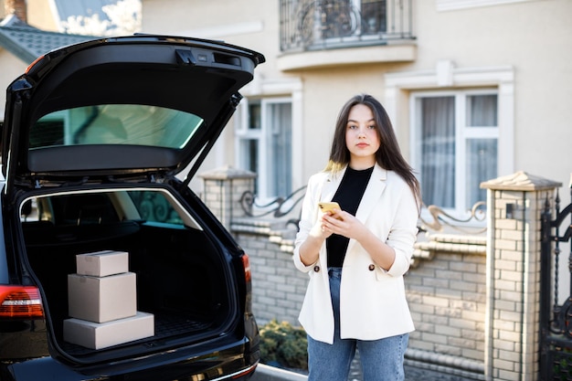 Młoda biznesowa kobieta stojąca przy samochodzie z paczkami wracającymi do domu samochodem Dziewczyna stoi w pobliżu samochodu i trzyma telefon komórkowy Koncepcja kupowania towarów przez Internet i dostarczania ich do domu