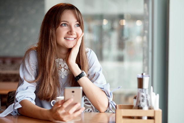 Młoda biznesowa kobieta używa jej mądrze telefon i ono uśmiecha się w sklep z kawą