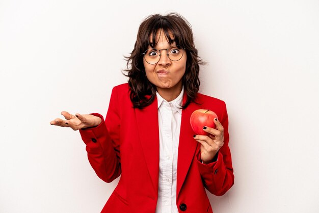 Młoda biznesowa latynoska kobieta trzymająca jabłko na białym tle wzrusza ramionami i otwiera oczy zdezorientowana