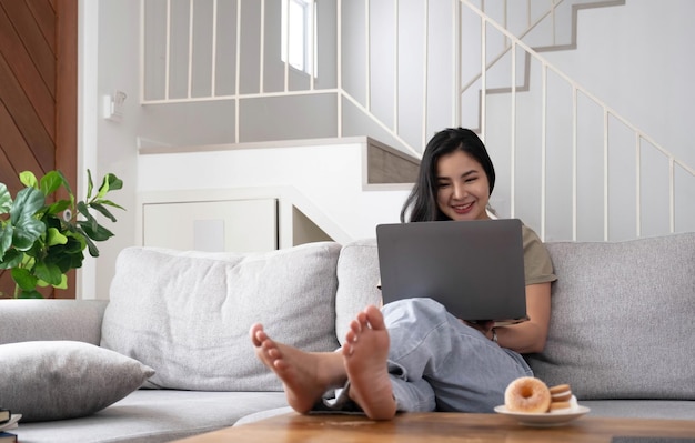 Młoda biznesowa niezależna Azjatycka kobieta pracująca na laptopie sprawdzająca media społecznościowe leżąc na kanapie podczas relaksu w salonie w domu