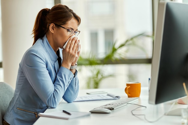 Młoda bizneswoman ma wirusa grypy i używa chusteczki podczas dmuchania nosa w biurze