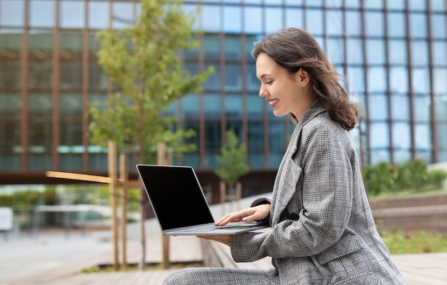 Młoda bizneswoman przeglądająca stronę internetową na laptopie siedząca na zewnątrz widok boczny