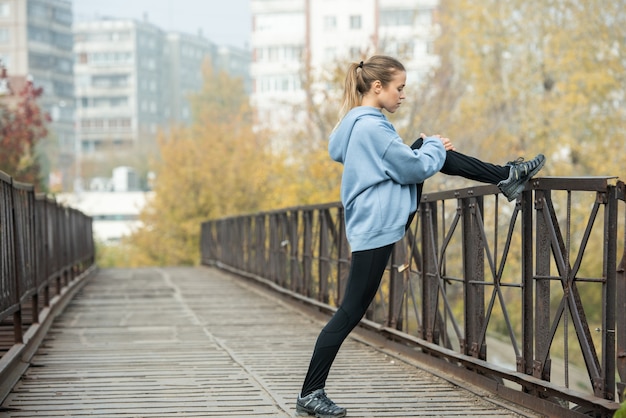 Młoda blond aktywna kobieta w leginsach i bluzie z kapturem stojąca na moście z lewą nogą zgiętą w kolanie na metalowym ogrodzeniu i ćwicząca rano