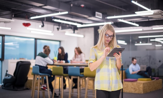 młoda blond bizneswoman pracująca online przy użyciu cyfrowego tabletu, stojąc w nowoczesnym biurze startowym z wieloetnicznym zespołem biznesowym w tle