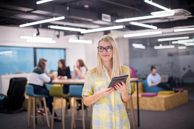 młoda blond bizneswoman pracująca online przy użyciu cyfrowego tabletu, stojąc w nowoczesnym biurze startowym z wieloetnicznym zespołem biznesowym w tle