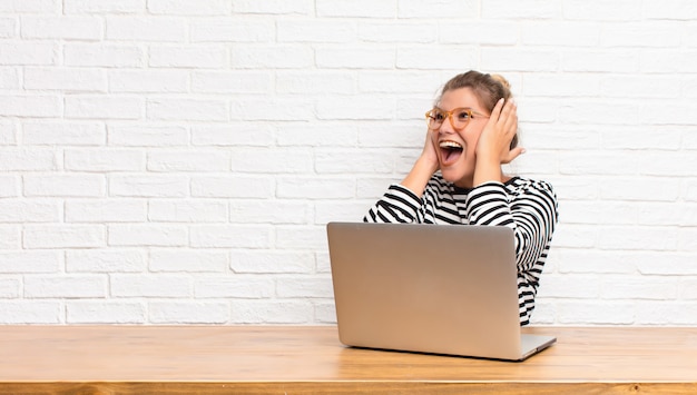 Młoda blond kobieta czuje się szczęśliwa, podekscytowana i zaskoczona, patrząc z boku obiema rękami na twarzy siedzącej z laptopem