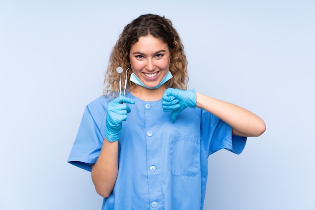 Młoda blond kobieta dentysta trzymając narzędzia na niebieskim tle dumny i zadowolony z siebie